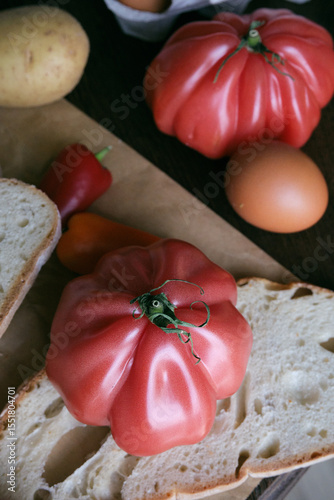 A rustic flat lay featuring slices of homemade artisan bread, fresh brown eggs in a carton, heirloom tomato, sweet peppers, and potatoes on a wooden kitchen table. Natural light and cozy.
