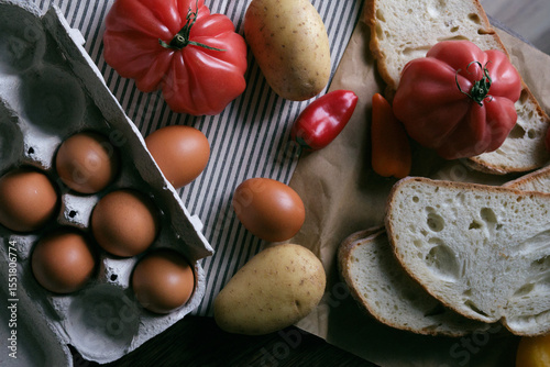 A rustic flat lay featuring slices of homemade artisan bread, fresh brown eggs in a carton, heirloom tomato, sweet peppers, and potatoes on a wooden kitchen table. Natural light and cozy.