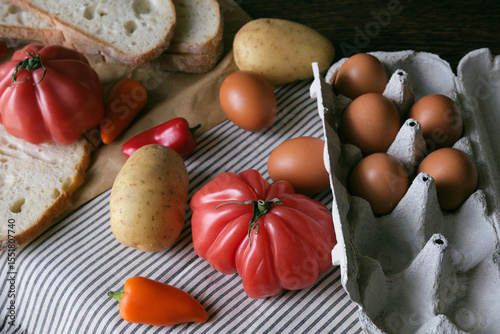 A rustic flat lay featuring slices of homemade artisan bread, fresh brown eggs in a carton, heirloom tomato, sweet peppers, and potatoes on a wooden kitchen table. Natural light and cozy.