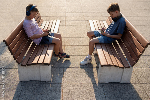 Children playing mobile game on bench in sunset light, focused on phone screen. Two boys using smartphones, watching stream, gaming, learning content outdoors. Digital childhood, education, technology