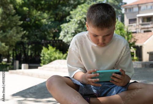 Little boy play online video game on smartphone while sitting in park. Child using phone looking at screen alone outside. Children and technology, education, lifestyle concept