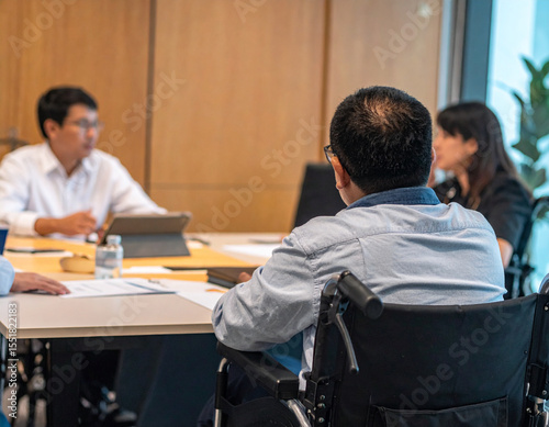 Professional documentary-style photo of wheelchair user collaborating with colleagues at modern office desk, natural office lighting through large windows, authentic teamwork interaction, accessible w