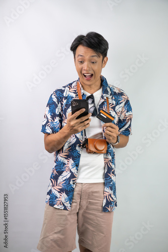 Handsome young asian man wearing blue summer shirt is holding smartphone with blank screen and dummy credit card, isolated over white background.