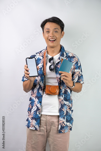 Handsome young asian man wearing blue summer shirt is holding smartphone with blank screen and indonesian passport, isolated over white background.