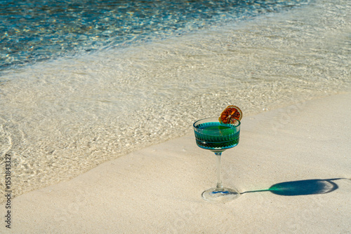 A clear, blue-toned cocktail, with a dried blood orange slice on the rim of the glass as a decoration. The glass is placed on the edge of a white sandy beach.