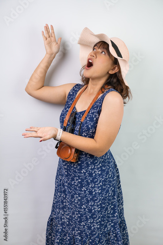 Happy beautiful young asian woman wearing blue summer dress and beach straw hat is pointing using index finger a copy space above her, isolated over white background