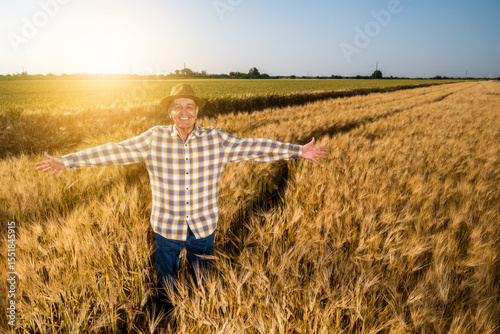 Happy senior man is standing in his growing wheat field in sunset. Farmer is satisfied with growth of crops. Portrait of happy senior man in wheat field.