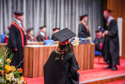 Back view of a female graduate walking on stage to receive her diploma during a graduation ceremony. She is wearing a traditional graduation gown and a square academic cap known as a mortarboard