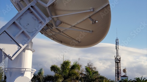 Large radio telescope searching for extraterrestrial signals, with a telecommunications tower in the background and palm trees in the foreground under a cloudy sky