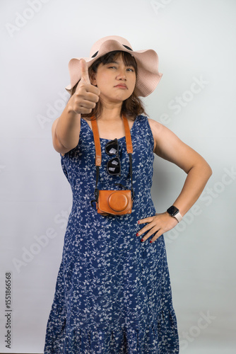 Happy beautiful young asian woman wearing blue summer dress and beach straw hat is showing thumbs up hand gesture, isolated over white background.