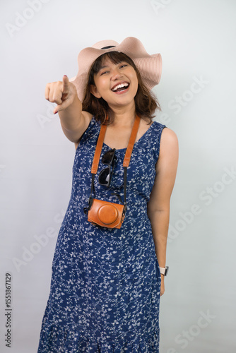 Happy beautiful young asian woman wearing blue summer dress and beach straw hat is pointing to the camera using her index finger, isolated over white background.