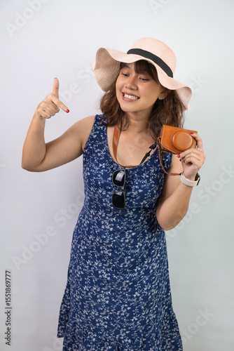 Happy beautiful young asian woman wearing blue summer dress and beach straw hat is pointing a copy space below her, isolated over white background.