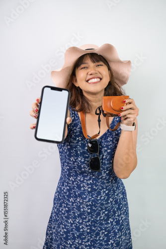 Happy beautiful young asian woman wearing blue summer dress and beach straw hat is holding a smartphone with blank screen for mock up mockup, isolated over white background