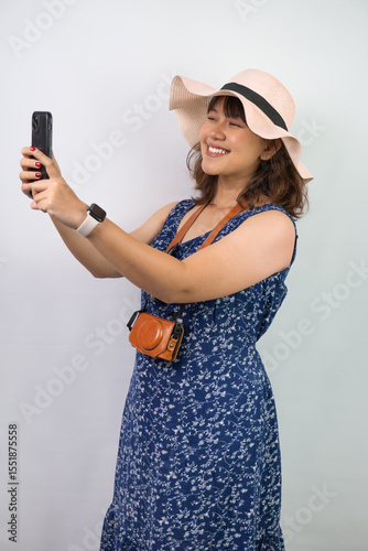 Beautiful young asian woman wearing blue summer dress and beach straw hat is posing while taking selfie photo using her smartphone, isolated over white background.