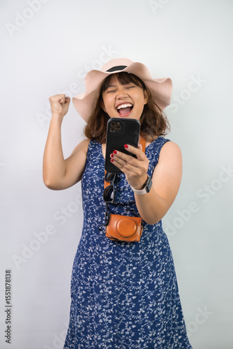 Happy beautiful young asian woman wearing blue summer dress and beach straw hat is holding a smartphone with blank screen for mock up mockup, isolated over white background