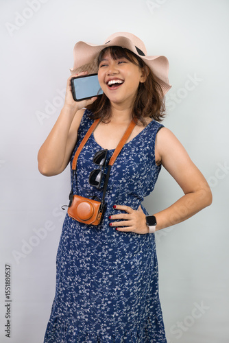 Beautiful young asian woman wearing blue summer dress and beach straw hat is talking with friend using smartphone with blank screen, isolated over white background.