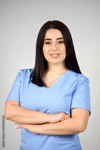 A cheerful, smiling female nurse in a blue medical suit on a white background. Portrait of a young nurse with long dark hair. The image conveys professionalism, compassion, and expertise in healthcare