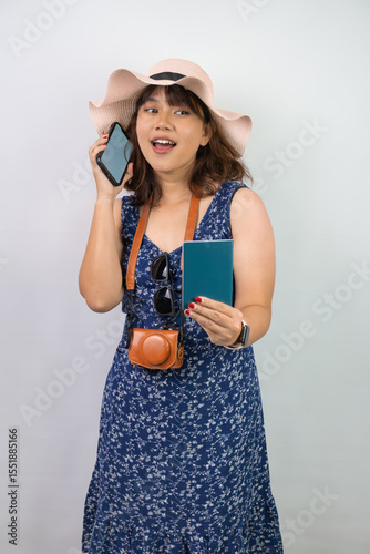 Beautiful young asian woman wearing blue summer dress and beach straw hat is holding smartphone with blank screen and indonesian passport, isolated over white background.