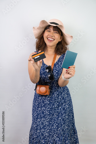 Beautiful young asian woman wearing blue summer dress and beach straw hat is holding indonesian passport and dummy credit card, isolated over white background.