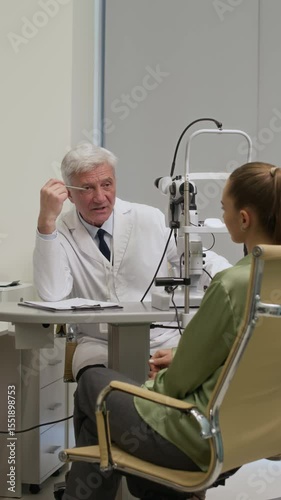 Vertical shot of mature male ophthalmologist talking to female patient during eye checkup appointment while prescribing glasses with specific lenes at doctors office in eye care center