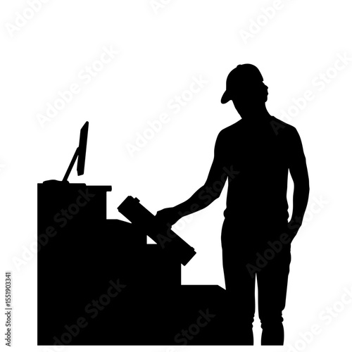 Silhouette of man wearing cap standing at table holding rolled up item with monitor on tabletop; suggests working, preparing or thinking in minimalist black and white composition.