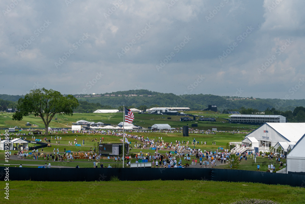 Obraz premium A large golf course is filled with spectators and event tents under a cloudy sky. An American flag stands prominently amidst the crowd, with distant hills in the background.