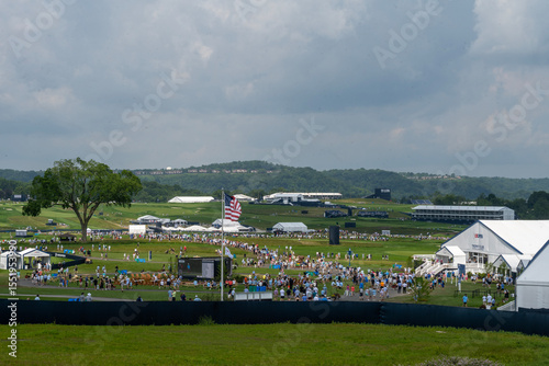 A large golf course is filled with spectators and event tents under a cloudy sky. An American flag stands prominently amidst the crowd, with distant hills in the background.