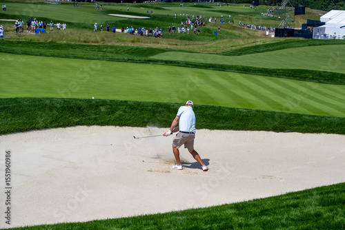  A golfer, captured in mid-swing, hits a golf ball from a sand bunker, sending sand flying. The dynamic shot is set against a lush green golf course.