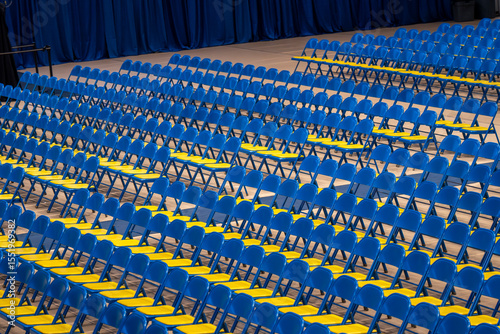 Rows of blue and yellow folding chairs are neatly arranged on a flat surface, creating a distinct pattern. The empty seating suggests preparation for an upcoming event or gathering.