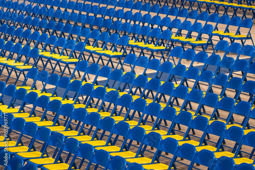 Rows of blue and yellow folding chairs are neatly arranged on a flat surface, creating a distinct pattern. The empty seating suggests preparation for an upcoming event or gathering.