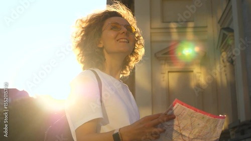 Backlit tourist with curly hair and yellow glasses, wearing a white t shirt and a backpack, smiles while holding a map in her hands in front of a historical building