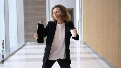 Happy businesswoman dancing in office corridor while holding tablet and coffee, enjoying work, celebrating success, or simply having fun during break