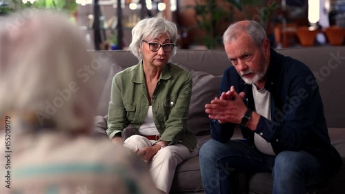 Seasoned senior couple sitting closely on living room sofa, intently sharing perspectives while using expressive hand movements during serious family discussion