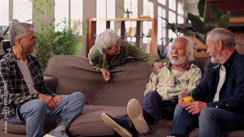 Happy senior friends, two men and a woman, are sitting on a comfortable sofa, engaging in lively conversation and enjoying refreshing drinks, creating a warm and joyful atmosphere