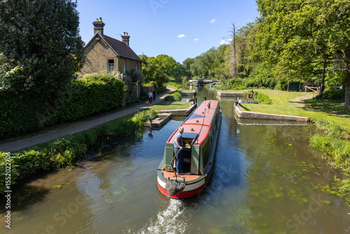 Canal barge being steered slowly through Walsham Gates Lock on the River Wey Navigation, while paddle boarders wait for it to pass