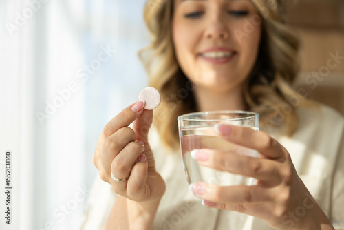 A smiling woman prepares to take a tablet with a glass of water in a bright, airy setting, emphasizing health and wellness.
