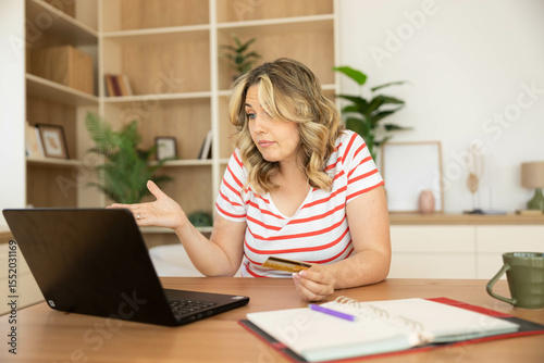 A young woman looks concerned while interacting with her laptop, holding a credit card in her hand, contemplating her purchase.