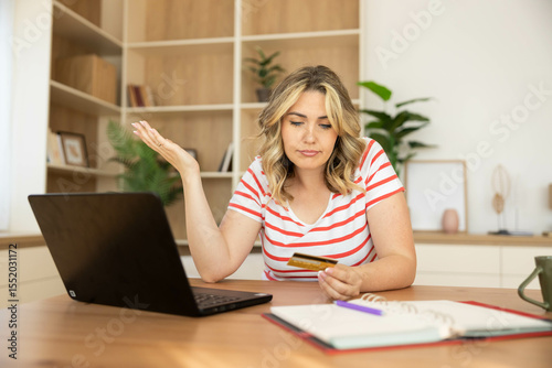 A young woman appears frustrated while trying to pay online, holding a credit card in front of her laptop.