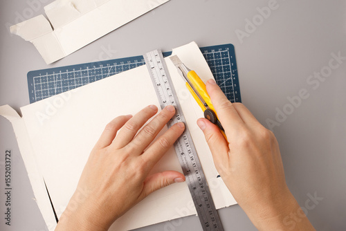 person cutting white cardboard with yellow utility knife and metal ruler on blue grid mat ideal for DIY projects, craft businesses, model making, creative workshops, precision cutting tools, hobby 