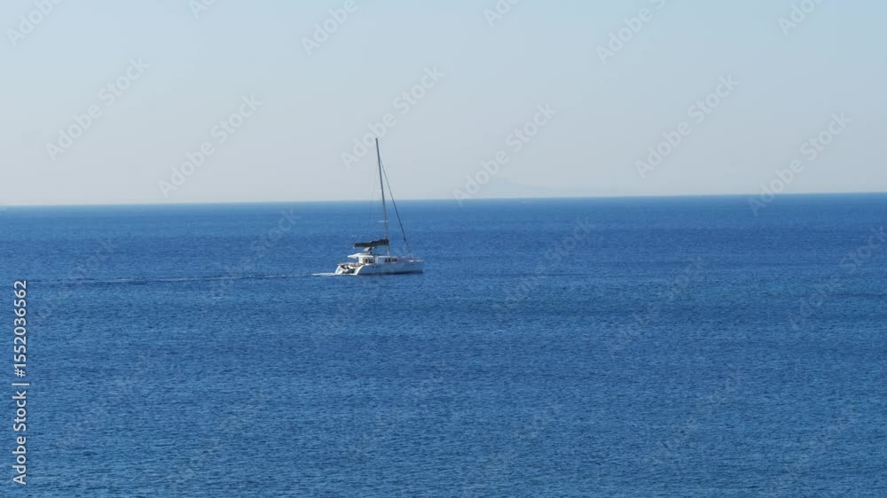 A white catamaran with dark mast sails moves across the calm, deep blue sea under a clear sky near Athens, Greece. The horizon shows a faint island outline, peaceful sailing conditions A white catamaran with dark mast sails moves across the calm, deep blue sea under a clear sky near Athens, Greece. The horizon shows a faint island outline, peaceful sailing conditions
