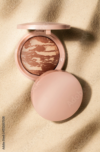 A makeup compact sits open on a textured sandy background, revealing a marbled bronzer with warm hues. The soft design emphasizes elegance and beauty.