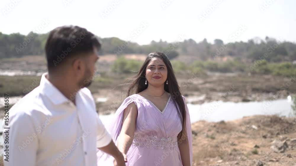 A cheerful Indian couple enjoys a sunny day at the beach, holding hands while walking along the shoreline. The woman is dressed in a casual maroon dress, and the man wears a white t-shirt with grey sh A cheerful Indian couple enjoys a sunny day at the beach, holding hands while walking along the shoreline. The woman is dressed in a casual maroon dress, and the man wears a white t-shirt with grey sh