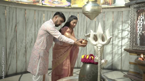 An Indian couple performs the sacred Abhishekam ritual to a Shiva Lingam in a Hindu temple. The woman is dressed in an elegant silk saree with jasmine flowers in her hair, and the man wears traditiona