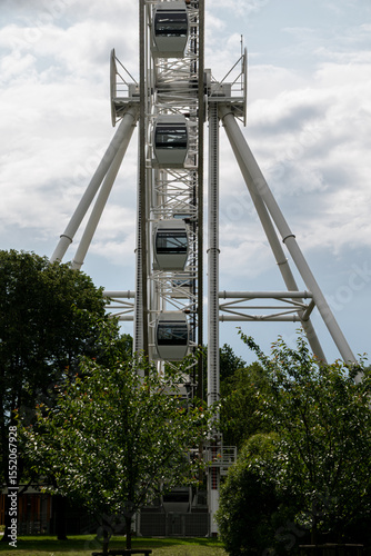 Modern white Ferris wheel with enclosed cabins viewed from the front, surrounded by green trees and cloudy sky in the background