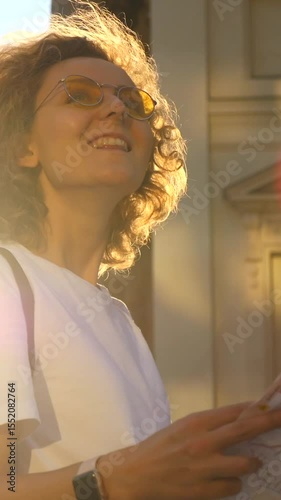 Vertical screen backlit curly haired female traveler wearing yellow sunglasses, holding city map, smiling cheerfully during golden hour sunset with urban landscape background