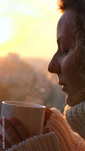 Vertical screen warm sunlight illuminates a woman's serene profile as she savors a hot drink, enveloped in a cozy sweater, against the backdrop of a breathtaking cityscape at dawn