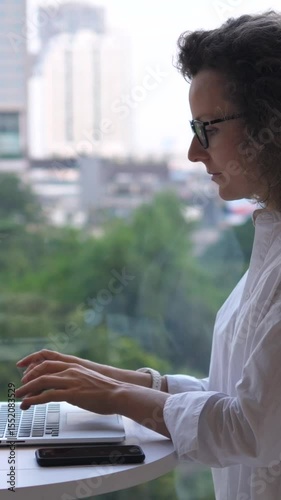 Vertical screen businesswoman adjusts glasses, types on laptop, and rests hands on keyboard while standing at a table near a large window overlooking a cityscape with green spaces