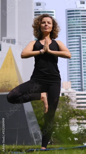 Vertical screen young woman with curly hair, dressed in sportswear, maintains balance in a tree pose, demonstrating body control and mindfulness against backdrop of modern city buildings