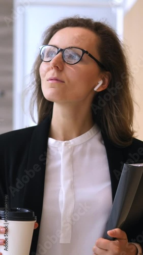 Vertical screen confident businesswoman walks through a modern office building, holding a takeaway coffee cup and a folder while wearing wireless earbuds and glasses