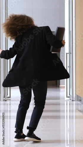 Vertical screen female professional dancing spontaneously in bright office hallway, holding coffee and files while expressing boundless energy and workplace joy with dynamic, carefree movement